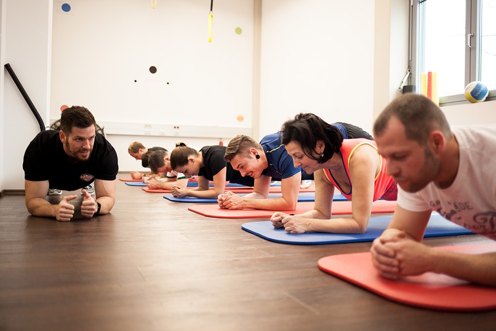 Trainer zeigt im Trainingsraum eine Armstütz-Übung auf dem Holzboden; sechs Patienten auf roten und blauen Matten folgen der gleichen Bewegung.