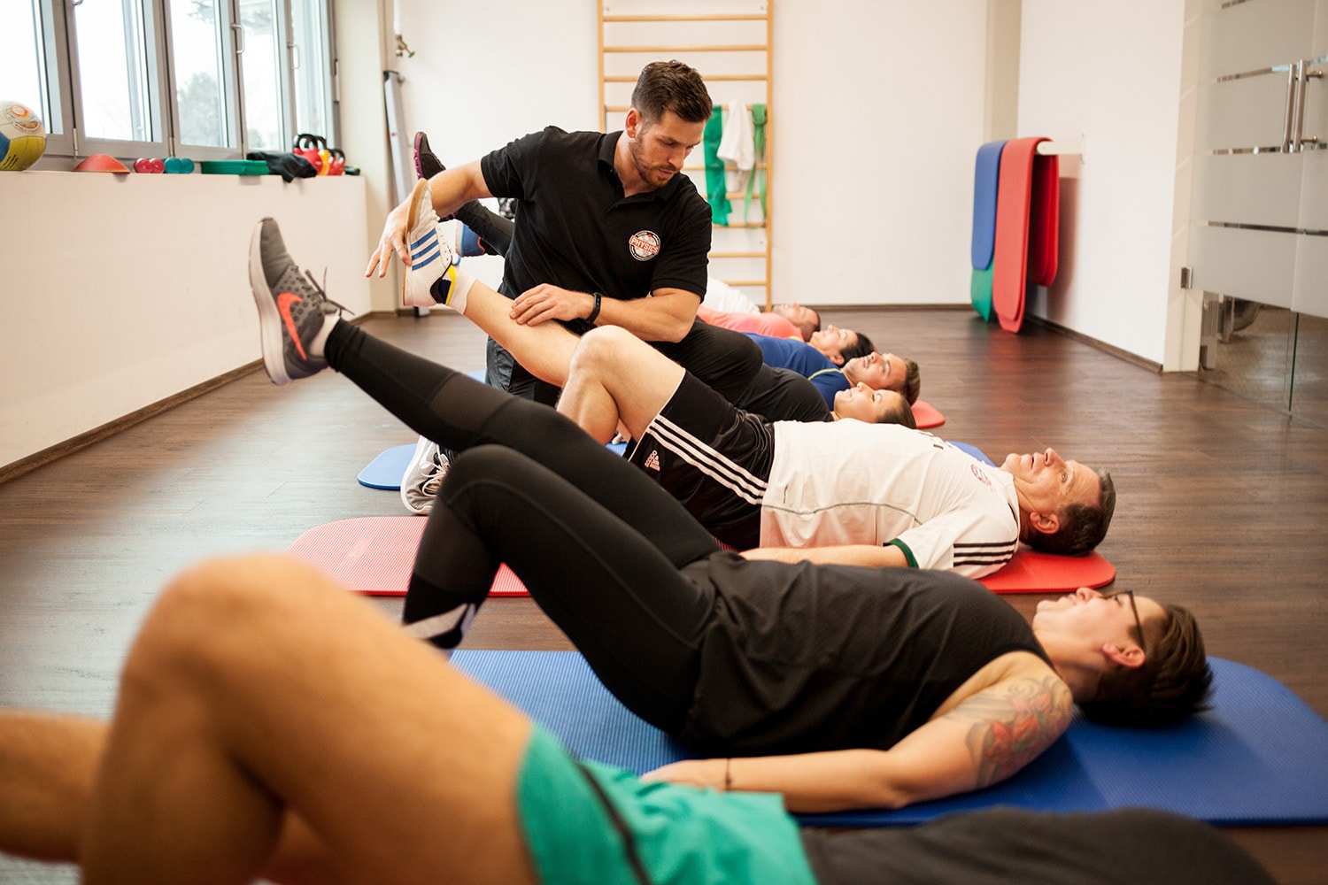 Mehrere Personen liegen im Trainingsraum auf dem Rücken und führen eine Übung mit angewinkeltem linkem Bein und gestrecktem, angehobenem rechtem Bein aus. Ein Physiotherapeut kniet neben einem Teilnehmer und unterstützt dessen Bein bei der korrekten Ausführung.
