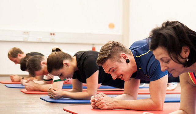Trainer zeigt im Trainingsraum eine Armstütz-Übung auf dem Holzboden; sechs Patienten auf roten und blauen Matten folgen der gleichen Bewegung.