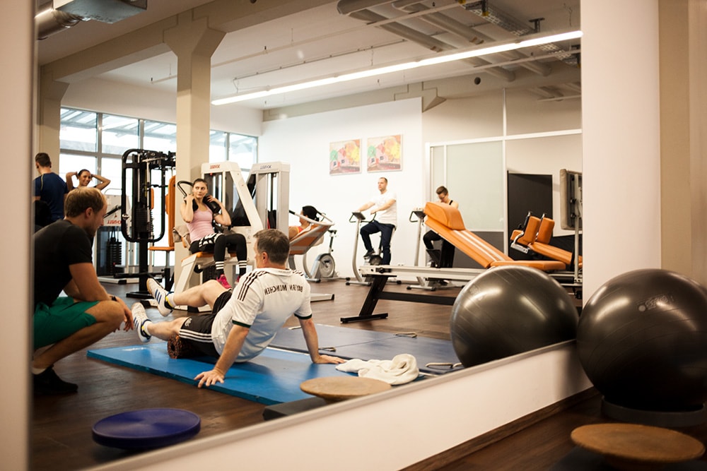 Großer Spiegel im Trainingsraum von PHYSICS Physiotherapie in Wasserburg, der Personen bei Übungen an Geräten, auf dem Stepper und am Boden sowie einen Trainer reflektiert.