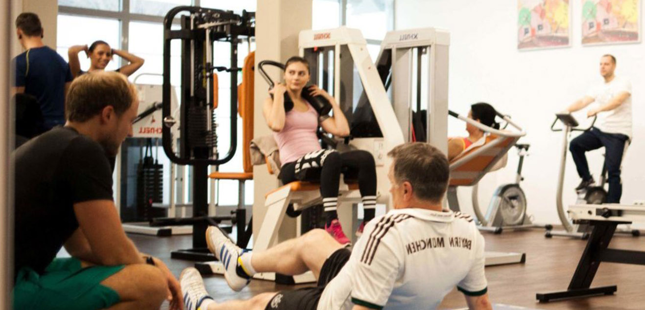 Großer Spiegel im Trainingsraum von PHYSICS Physiotherapie in Wasserburg, der Personen bei Übungen an Geräten, auf dem Stepper und am Boden sowie einen Trainer reflektiert.