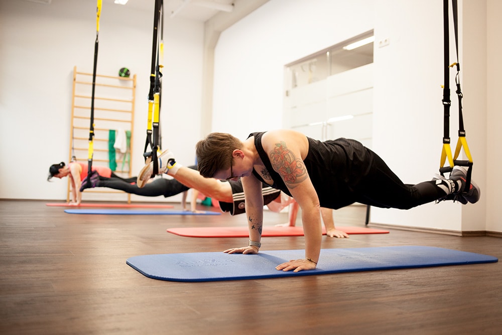 Eine Frau trainiert im Vordergrund im TRX-Liegestütz mit den Füßen in den Bändern, während im Hintergrund weitere Teilnehmer das TRX-Workout in derselben Übung ausführen.