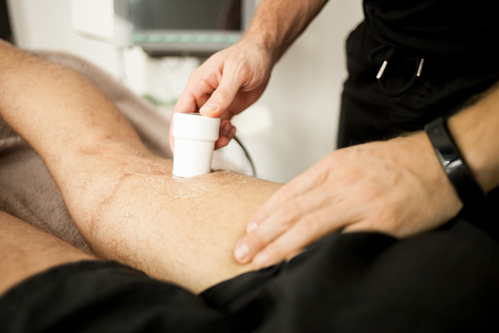 Bild auf dem ein Physiotherapeut mit einem Ultraschallgerät den hinteren Obershenkel eines liegenden Patienten untersucht