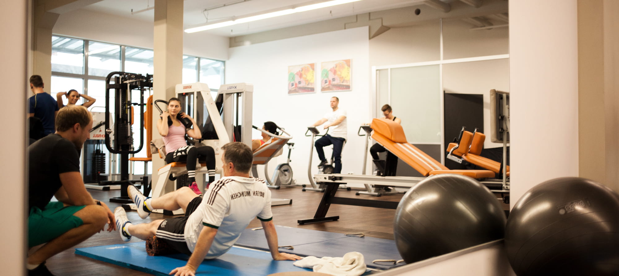 Großer Spiegel im Trainingsraum von PHYSICS Physiotherapie in Wasserburg, in dem sich ein schwarzer Trainingsball und mehrere Personen bei Übungen an Geräten, Steppern und am Boden sowie ein Trainer widerspiegeln.