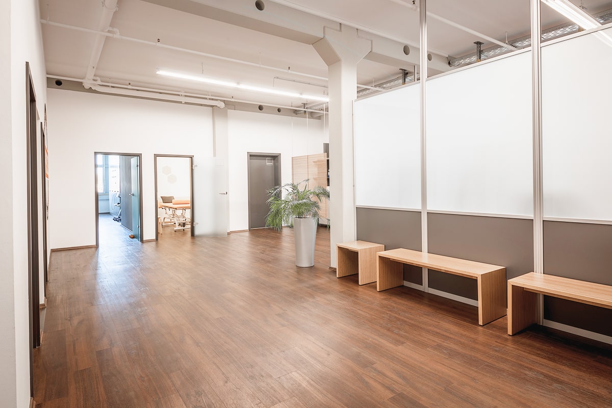 Wartebereich der Physiotherapie PHYSICS in Wasserburg am Inn, mit Holzbänken, Holzboden, einer Pflanze und halbtransparenten Trennwänden.